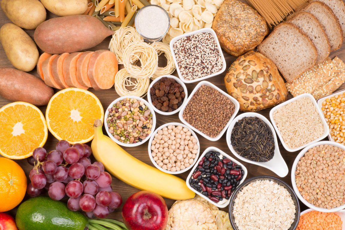Top-down view of carbohydrate-rich foods like sweet potatoes, whole grain bread, pasta, beans, lentils, bananas, and grapes.