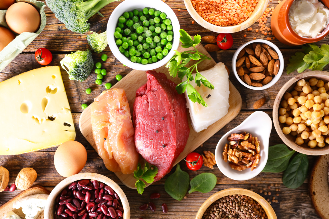 Selection of protein-rich foods including raw chicken, beef, fish, eggs, cheese, beans, lentils, and fresh broccoli.