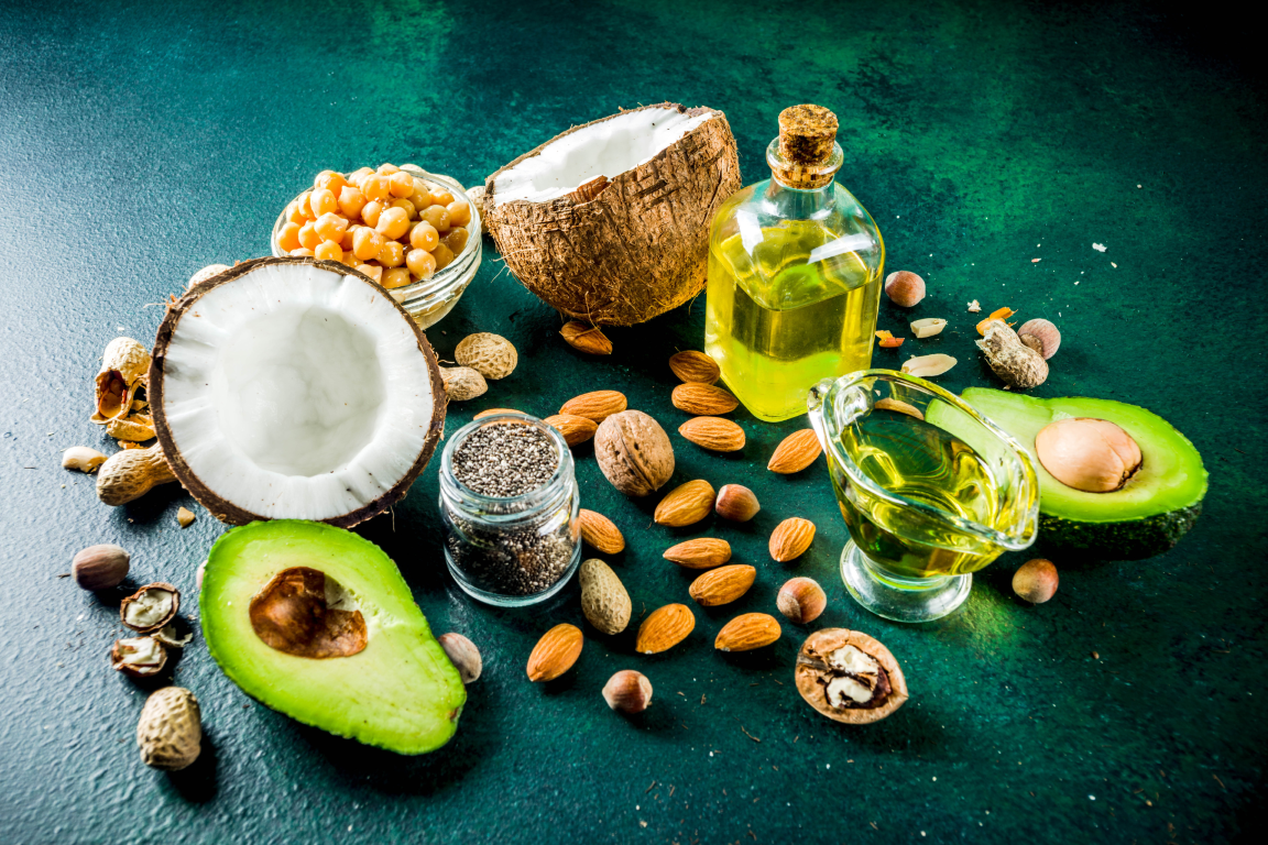 Assortment of healthy fats including sliced avocado, fresh coconut, walnuts, almonds, chia seeds, and olive oil bottles.