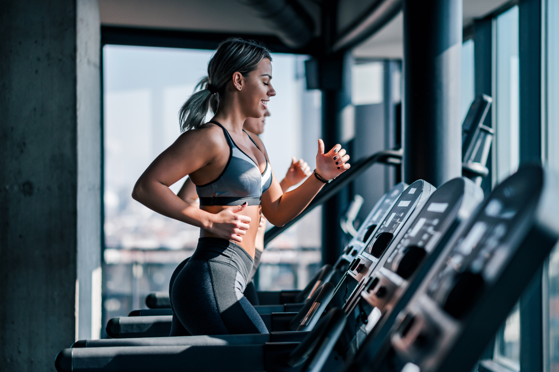 A smiling young woman performing cardio exercise on a treadmill in a modern gym with large windows in the background.
